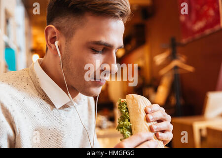 Photo de jeune homme séduisant habillé en chemise blanche assis dans le café tout en maintenant l'écoute et de sandwich de la musique. Banque D'Images