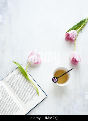 Nature morte avec trois tulipes perroquet ébouriffé, tasse de thé, cuillère à thé, livre, vue de dessus, le titre, sur fond gris et blanc Banque D'Images