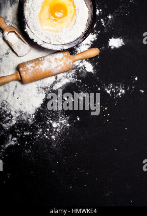 Ingrédients et ustensiles pour la préparation de produits de boulangerie - farine, la pâte à pain, œufs, rouleau à pâtisserie, fouetter, tamis, pain - sur table noire avec c Banque D'Images