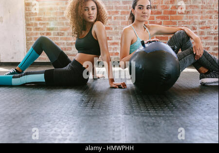 Portrait de deux jeunes femmes assises sur le plancher du gymnase avec ballon. Mixed Race femelles dans healthclub. Banque D'Images