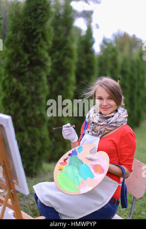 Mignon femelle peintre posant, riant et en face de l'appareil photo , attire et souriant avec des pinceaux et de la palette de couleurs à la main, est assis sur une chaise près de chevalet dans Banque D'Images