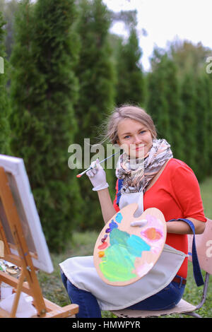 Mignon femelle peintre posant, riant et en face de l'appareil photo , attire et souriant avec des pinceaux et de la palette de couleurs à la main, est assis sur une chaise près de chevalet dans Banque D'Images