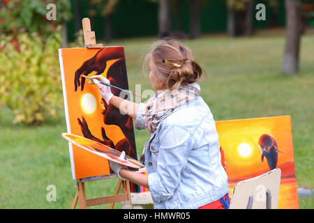 Jeune femme artiste termine la peinture qui montre femme qui ne cesse de soleil sur fond orange, assis le dos au photographe et derrière Banque D'Images