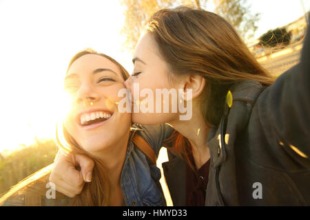 Deux amis affectueux baisers et prendre une à l'extérieur dans la rue selfies au coucher du soleil avec une lumière chaude dans l'arrière-plan Banque D'Images