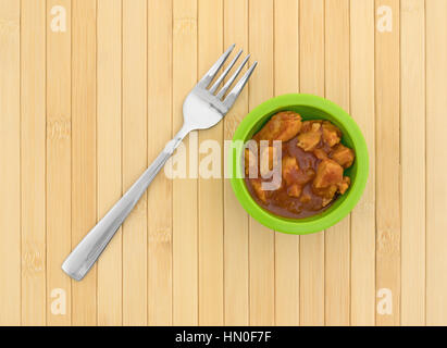 Un petit bol vert rempli de morceaux de poulet dans une sauce épaisse avec une fourchette pour le côté sur un napperon en bois. Banque D'Images