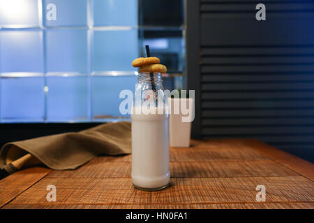 Le flacon en verre avec du lait et noir paille qui a mis deux cookies, chambre verte fleurs dans vase rectangulaire blanc et marsh drape dans ta carré en bois Banque D'Images