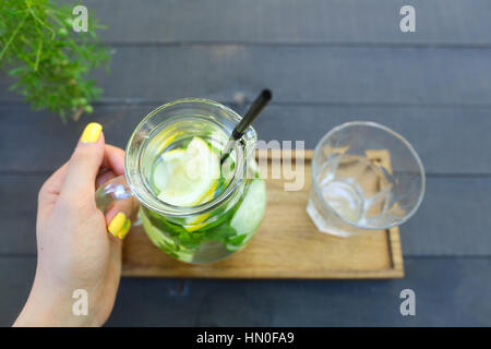 Carafe en verre qui est tenu en main avec verre de concombre, de citron, de menthe et de verre vide coupe à facettes se tient sur planche de bois sur gris woode Banque D'Images