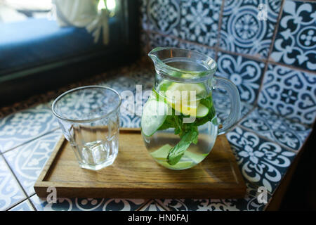 Carafe en verre avec verre de concombre, de citron, de menthe et de verre vide coupe à facettes se dresse sur le carrelage avec des modèles dans un café. Concept de refreshin Banque D'Images