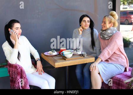 Trois jeunes jolies filles amies chatter, le commérage, partager des secrets, rire, sourire et assis sur une chaise à table et l'une des filles parle par pho Banque D'Images