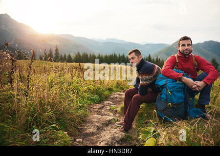 Deux jeunes hommes en matériel de randonnée assis sur un chemin au lever du soleil tout en prenant une pause de trekking dans le désert Banque D'Images