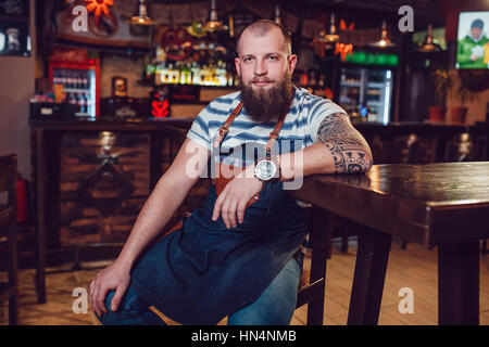 Barman barbu avec tatouages et wathes portant un tablier, assis dans un bar. Banque D'Images