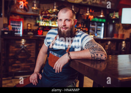 Barman barbu avec tatouages et wathes portant un tablier, assis dans un bar. Banque D'Images