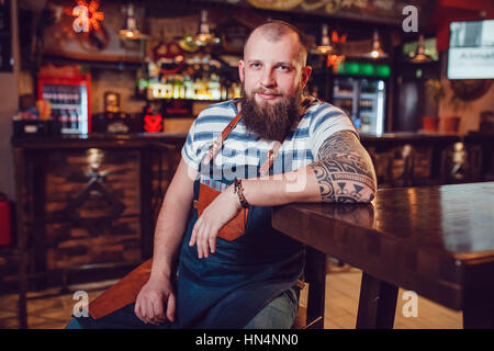 Barman barbu avec tatouages et wathes portant un tablier, assis dans un bar. Banque D'Images