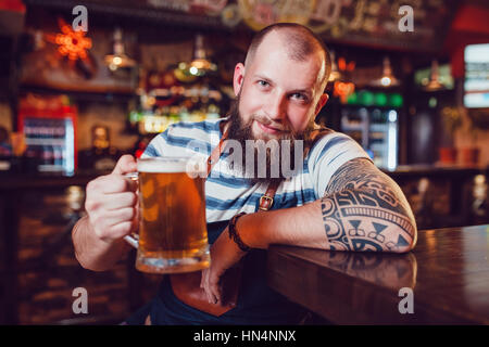 Barman barbu avec des tatouages portant un tablier, assis dans un bar et la tenue d'un verre de bière Banque D'Images