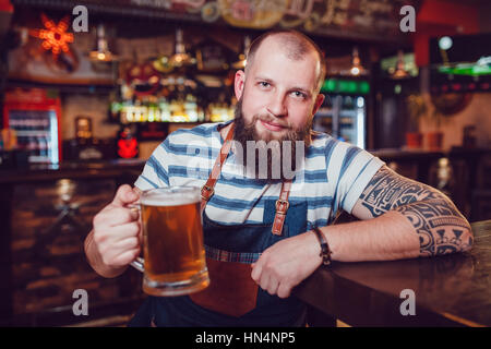Barman barbu avec des tatouages portant un tablier, assis dans un bar et la tenue d'un verre de bière Banque D'Images