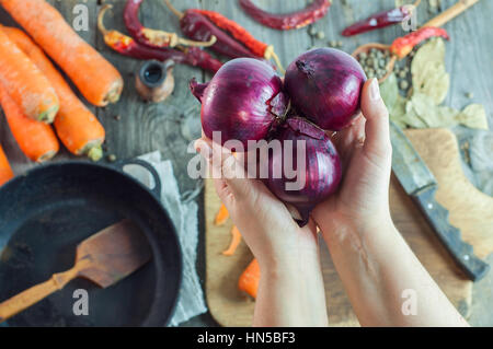 Les mains tenant trois oignons rouges sur le fond du tableau avec des légumes et épices, près de poêle en fonte noire avec une spatule en bois, haut v Banque D'Images