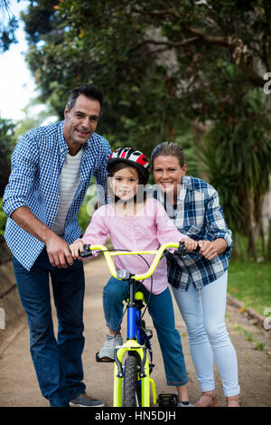 Portrait of happy parents aider fille à circuler à bicyclette dans le parc par une journée ensoleillée Banque D'Images