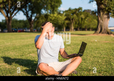 Jeune homme triste avec ordinateur portable et smartphone, assis dehors dans l'herbe verte du parc en plaçant la main sur la tête à cause du stress maux de tête et les yeux fermés à la d Banque D'Images
