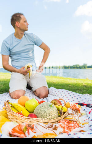 Jeune homme assis sur une couverture de pique-nique de la banane de l'alimentation par le panier de fruits à la rivière à Banque D'Images