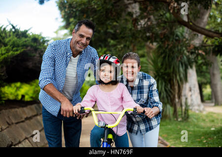 Portrait of happy parents aider fille à circuler à bicyclette dans le parc par une journée ensoleillée Banque D'Images