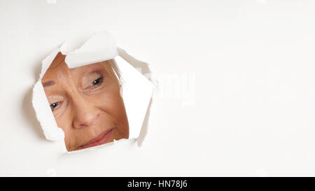Curieux femme plus âgée à la recherche à travers le trou dans du papier blanc déchiré au fond de l'espace de copie. Vue panoramique 16:9 format d'en-tête ou de bannière. Banque D'Images