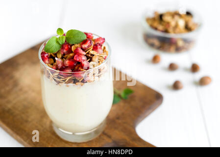 Petit-déjeuner sain concept. Crème fraîche vanille avec granola dans un verre garni de grenadier et menthe disposées sur une table en bois blanc. Banque D'Images
