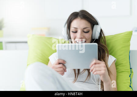 Girl with headphones relaxing on le lit à la maison et regarder des vidéos en ligne, elle est souriante et profiter de Banque D'Images