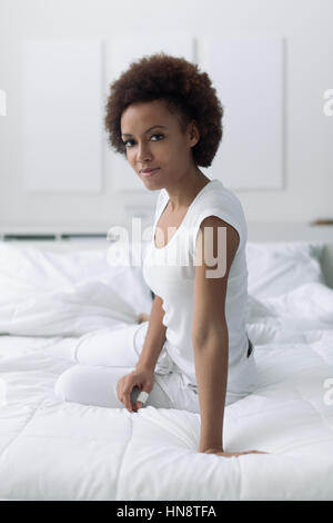 Young attractive african woman relaxing on le lit à la maison, elle est smiling at camera Banque D'Images