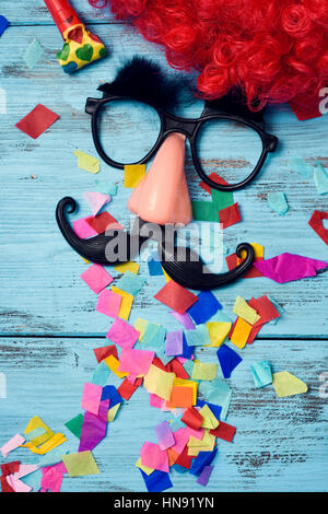 Une perruque rouge, une paire de lunettes noires avec de faux sourcils, un nez et une moustache formant la face d'un homme sur une surface en bois rustique bleu plein de confett Banque D'Images
