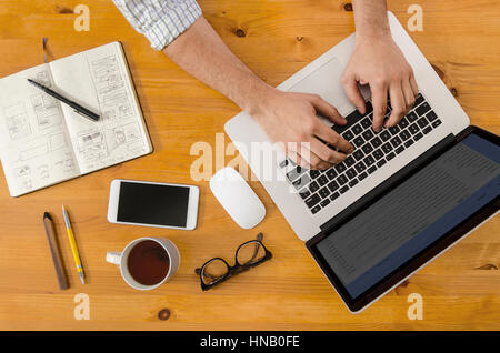 Mains de la saisie sur un ordinateur portable dans un environnement de travail de démarrage avec les outils de conception, l'ordinateur portable, stylo, et téléphone. Vue ci-dessus. Banque D'Images
