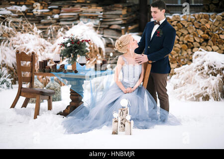 Mariage d'hiver sur la neige en plein air et le bois de l'arrière-plan. Mariée est assise, le marié se tient à proximité. À côté d'eux est le tableau avec des gobelets de vin et bouquet. Banque D'Images