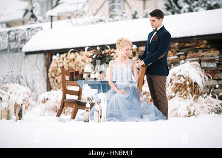 Mariage d'hiver en plein air sur fond de maison couverte de neige. Mariée est assise sur une chaise, le marié se trouve à proximité. À côté d'eux est président et de table avec des gobelets Banque D'Images