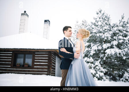 Mariage d'hiver en plein air sur fond de maison couverte de neige et de sapins. Mariée et le marié sont debout et s'étreindre. Banque D'Images