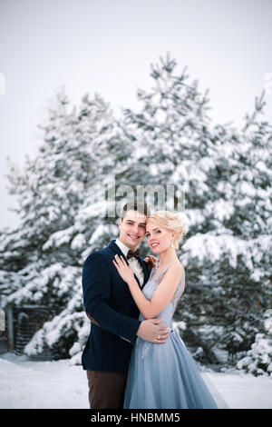 Mariage d'hiver en plein air sur fond de sapins couverts de neige. Mariée et le marié sont debout et s'étreindre. Loin est visible une grande première. Banque D'Images