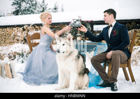 Mariée et le marié en hiver avec malamute chien assis à table et verres de vin. clink Sur la table est bouquet. Banque D'Images