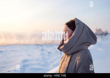 Jeune jolie fille pour le coucher du soleil, debout sur la berge d'une rivière gelée. Elle vêtue de beige veste avec une capuche Banque D'Images