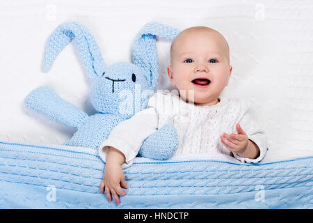 Drôle de petit bébé portant une veste en tricot chaud Playing with toy bunny détente sur le câble blanc Couverture tricot sous le soleil de pépinière. Les enfants et les vêtements d'hiver Banque D'Images