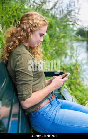 Young caucasian woman reads sur téléphone mobile à côté de l'eau banc Banque D'Images