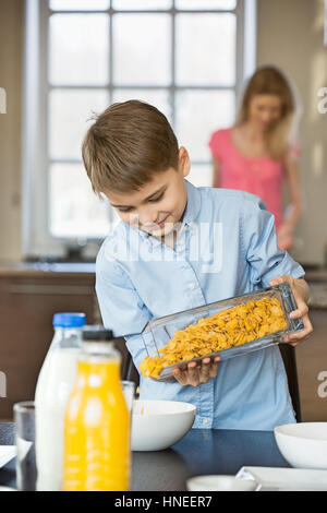 Boy pouring flocons de maïs dans un bol avec la mère debout en arrière-plan Banque D'Images