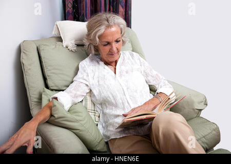 Senior woman reading book tout en vous relaxant sur un fauteuil à la maison Banque D'Images