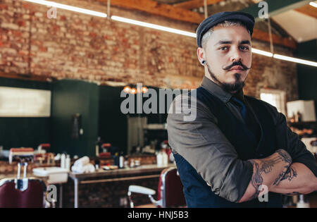 Portrait de jeune homme debout avec les bras croisés au salon de coiffure. Coiffure homme debout dans son salon de coiffure. Banque D'Images