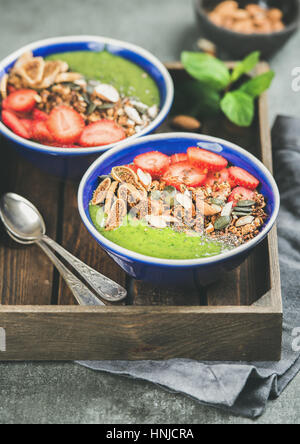 Petit déjeuner sain. Bols smoothie vert avec des fraises, granola, Chia et graines de citrouille, de fruits secs et d'écrou dans le bac en bois sur béton gris retour Banque D'Images