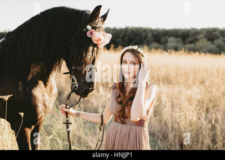 Portrait femme portant une robe beige flowy et couronne de fleurs, tenant les cordes de son cheval noir, regardant la caméra Banque D'Images