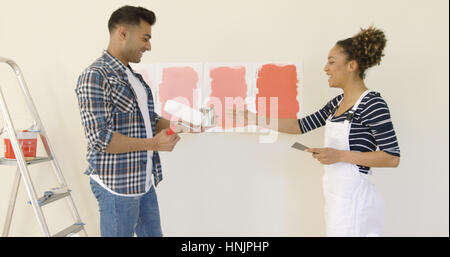 Laughing young couple choisir une couleur de peinture pour rénover leur nouvelle maison debout avec un rouleau et pinceau dans leurs mains en face d'échantillons sur le w Banque D'Images