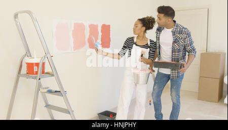 Jeune couple discuter de nuances de couleur de la peinture ensemble comme ils rénover leur appartement standing looking at quatre échantillons sur le mur Banque D'Images