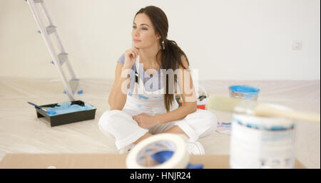 Jeune femme sérieuse rénovation d'une maison prendre une pause pour la décoration à s'asseoir sur le sol à la recherche au loin dans la distance avec une expression pensive Banque D'Images