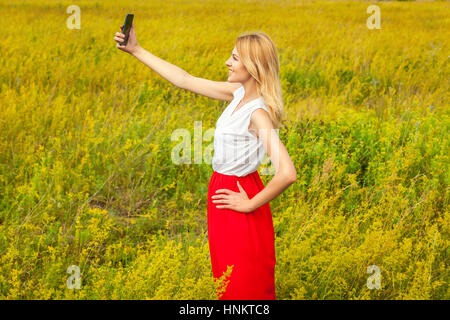 Blonde avec le téléphone. La belle fille blonde en robe rouge-blanc Montres téléphone mobile dans le domaine en vert. Banque D'Images