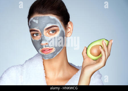 Jolie jeune fille tenant une demi-avocat en main. Photo Jeune femme avec masque d'argile sur fond bleu. Beauté et Soins De La Peau concept Banque D'Images