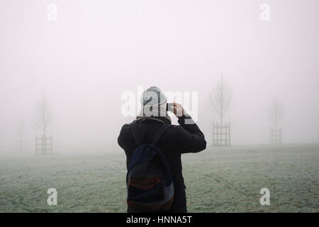 Jeune homme en manteau d'hiver, bonnet et écharpe en prenant des photos avec son téléphone portable de London park, disparaissant dans le brouillard sur froid matin d'hiver. Banque D'Images