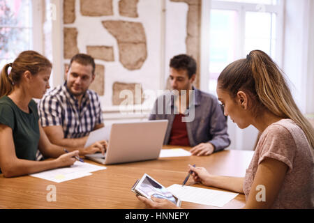 Jeune femme professionnelle contrôle ses notes sur sa tablette pendant que le reste de l'équipe sont à la recherche à un ordinateur portable à l'arrière-plan Banque D'Images